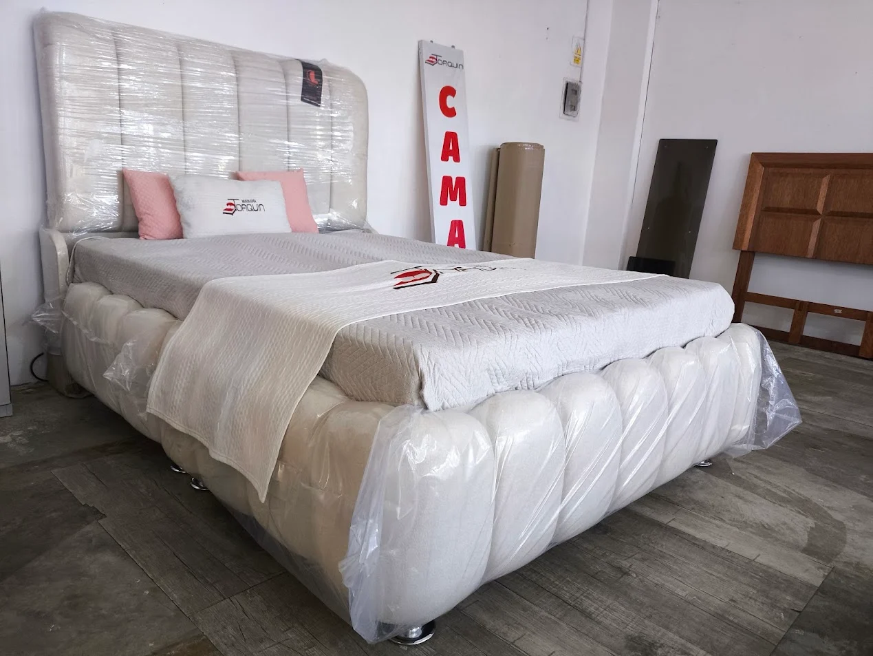 Cama tapizada de color claro fotografiada en el negocio, con cabecera acolchada y protección transparente.