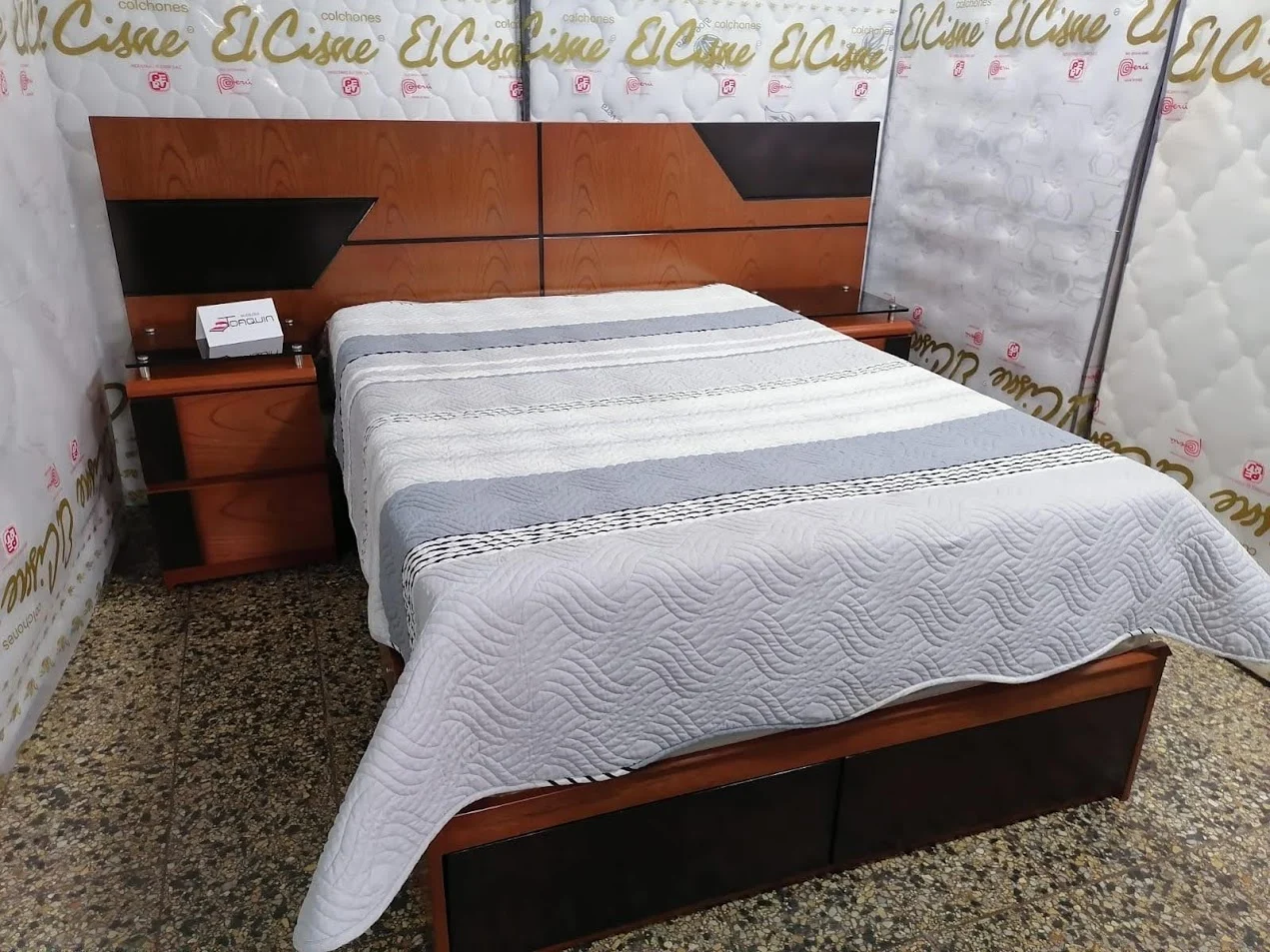 Cama del negocio con cabecera de paneles en tonos marrón y oscuro, veladores y ropa de cama clara.